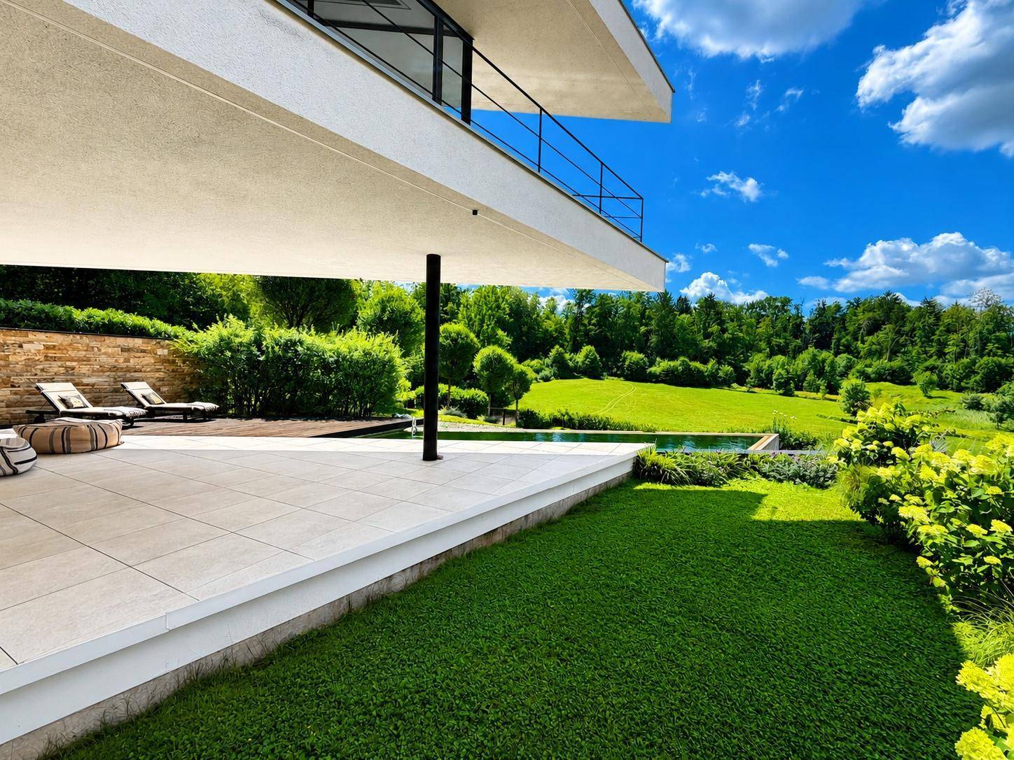 Gepflegter Gartenbereich mit Rasenfläche, Terrasse und Liegestühlen unter strahlend blauem Himmel.