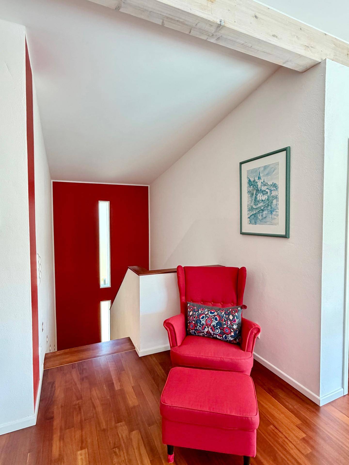 Heller Flur mit roter Akzentwand, Holzboden und gemütlichem Sessel als Leseecke.