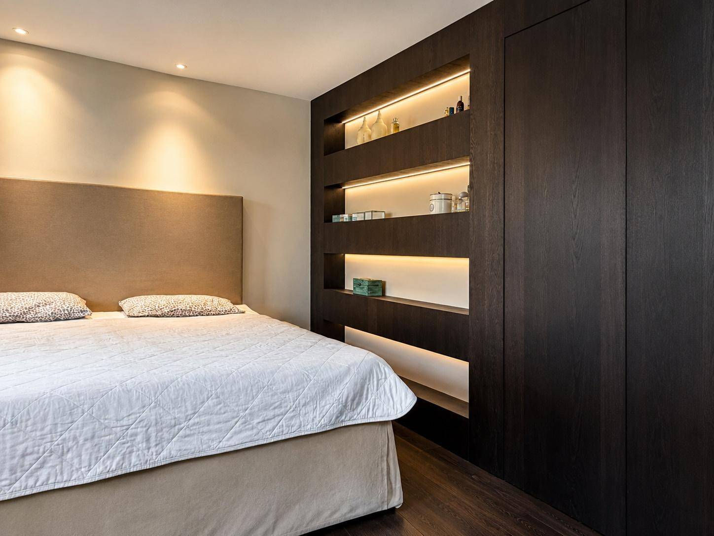 Modernes Schlafzimmer mit Einbauschränken, beleuchteten Regalen und dunklem Holzboden.