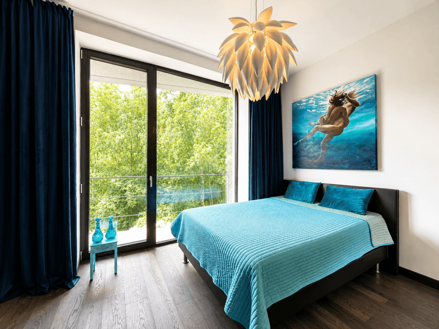 Helles Schlafzimmer mit großen Fenstern, Holzboden und blauer Dekoration mit Blick ins Grüne.