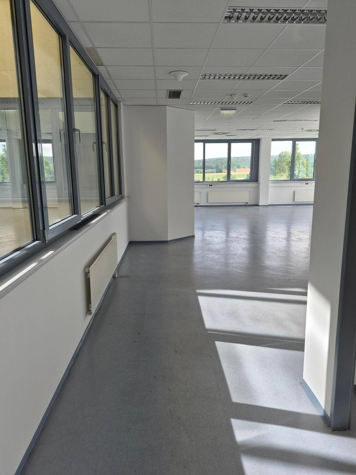 Langer, heller Bürobereich mit durchgehenden Fenstern und grauem Boden, der in weitere Räume führt.