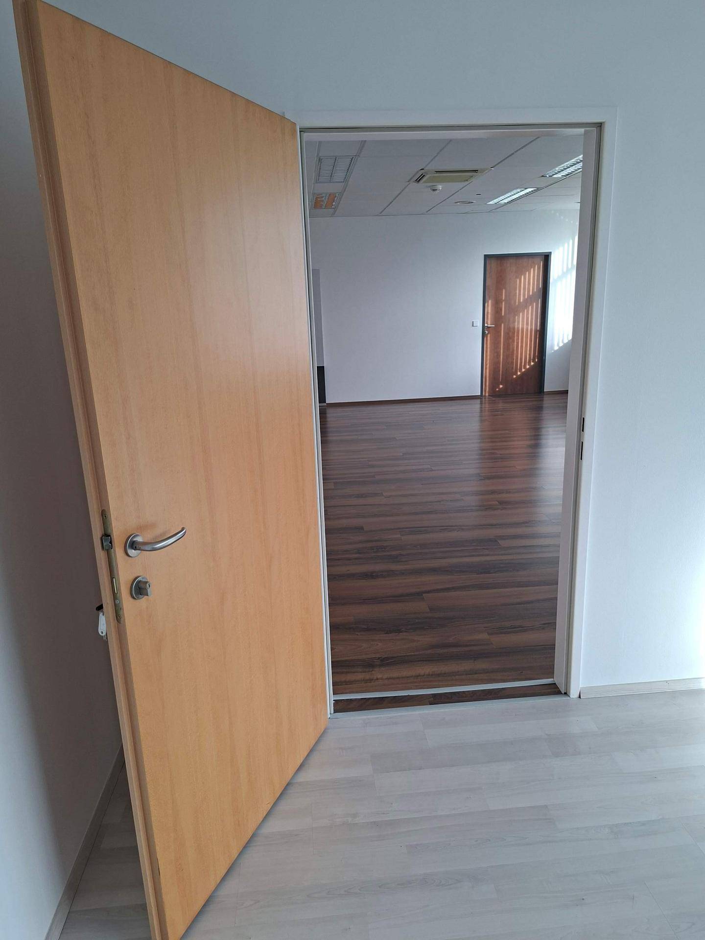 Offene Holztür, die den Übergang von hellem zu dunklem Laminatboden in angrenzenden Büroräumen zeigt.