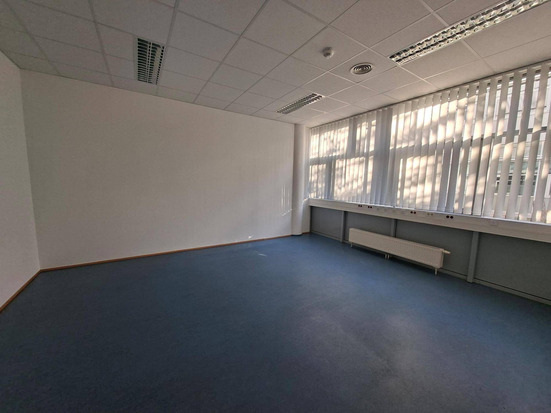 Heller Büroraum mit blauen Bodenfliesen und vertikalen Jalousien an den großen Fenstern.