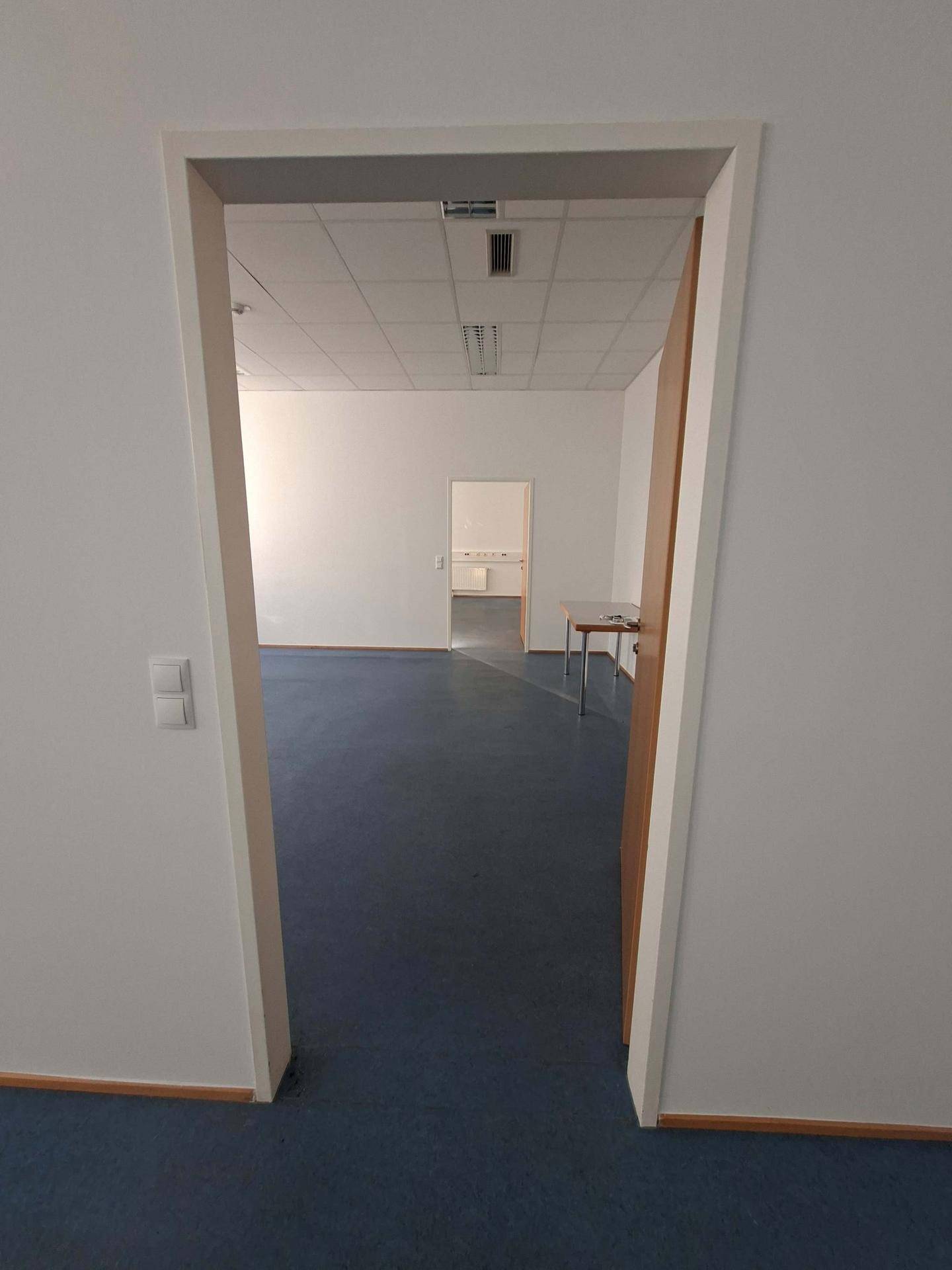 Blick durch eine offene Tür in einen weiteren Büroraum mit blauen Bodenfliesen und Deckenbeleuchtung.