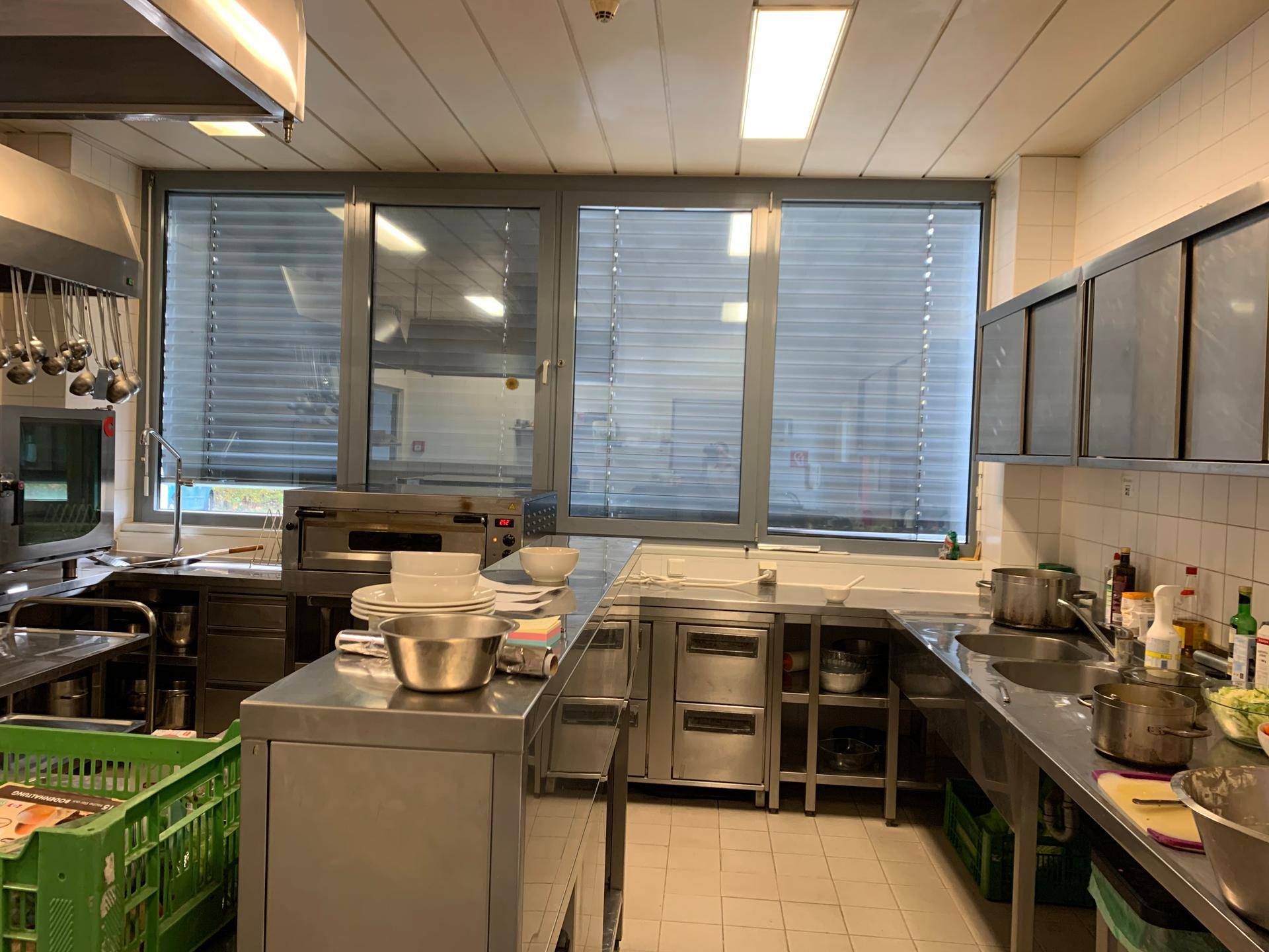 Funktionale Gewerbeküche mit Edelstahlarbeitsplatten, Spülbecken und großen Fenstern für ausreichend Licht.