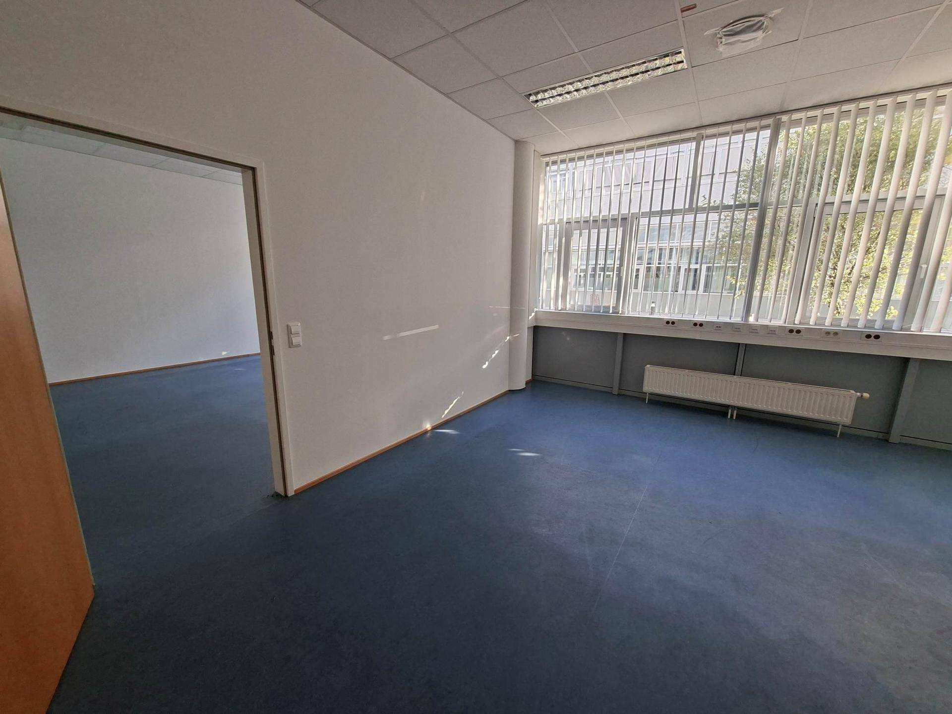 Geräumiger Büroraum mit blauen Bodenfliesen und Zugang zu einem weiteren Zimmer durch eine offene Tür.