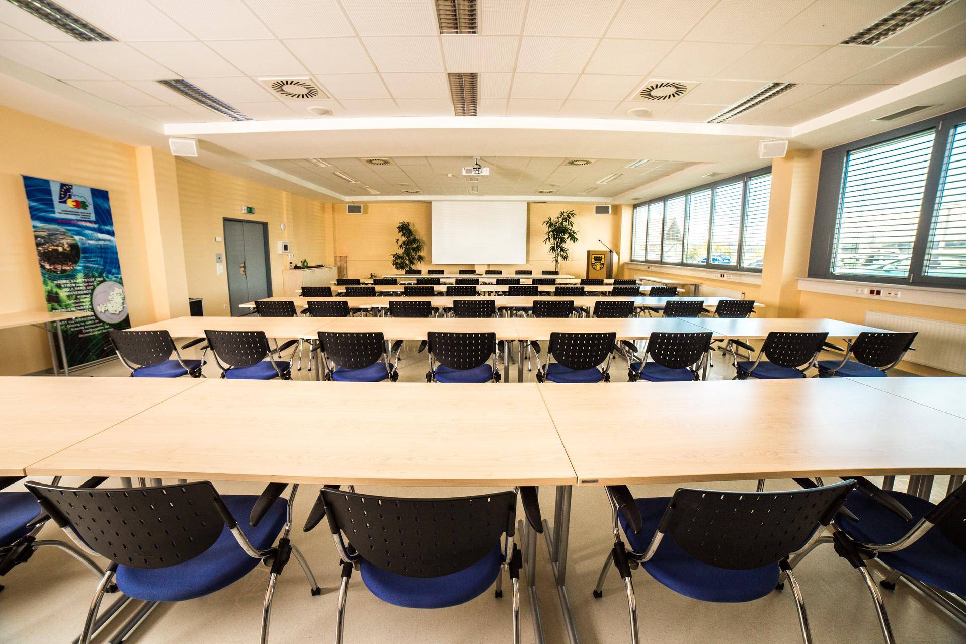 Großer Veranstaltungsraum mit Reihen von Tischen und Stühlen sowie einem Projektor für Präsentationen.