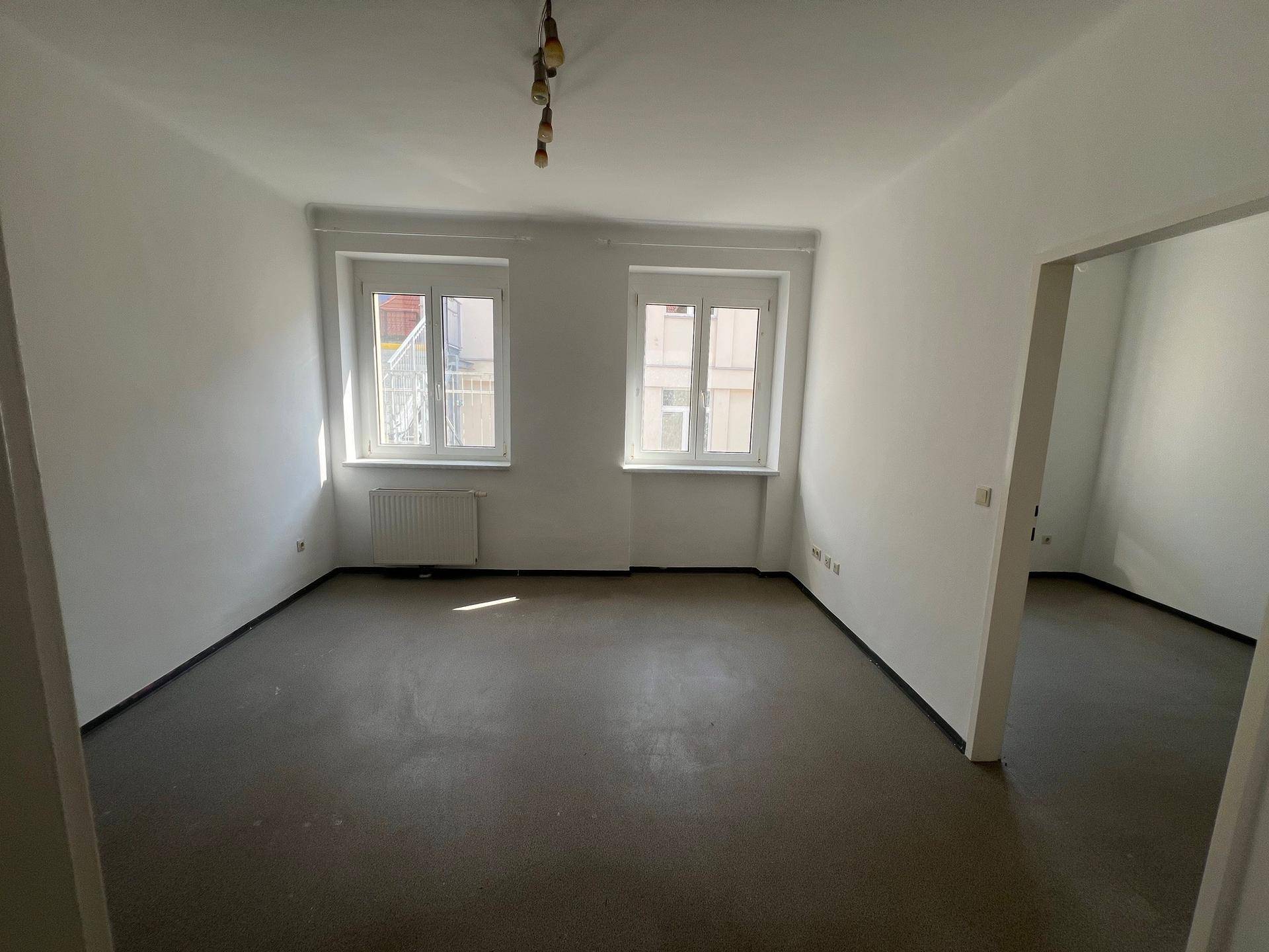 Helles, leeres Wohnzimmer mit zwei Fenstern, weißen Wänden und dunklem Boden, bereit zur individuellen Gestaltung.