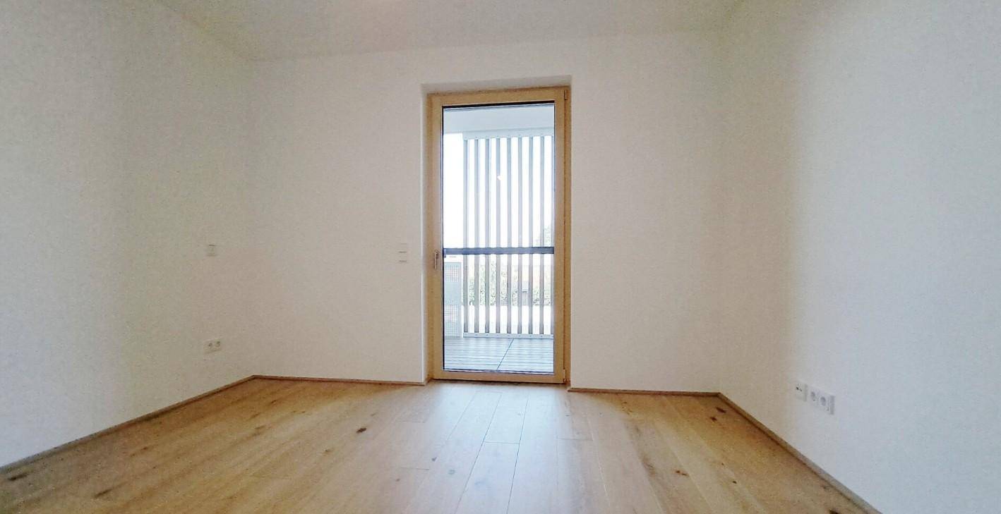 Unmöbliertes Zimmer mit Holzboden und einer Balkontür, die für viel Helligkeit sorgt.