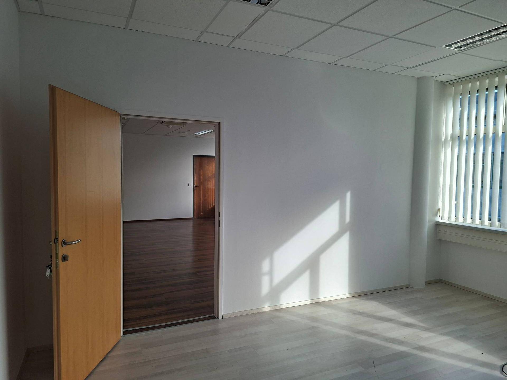 Leerer Büroraum mit hellem Laminatboden, offener Holztür und Fenster mit Jalousien, durch das Sonnenlicht fällt.