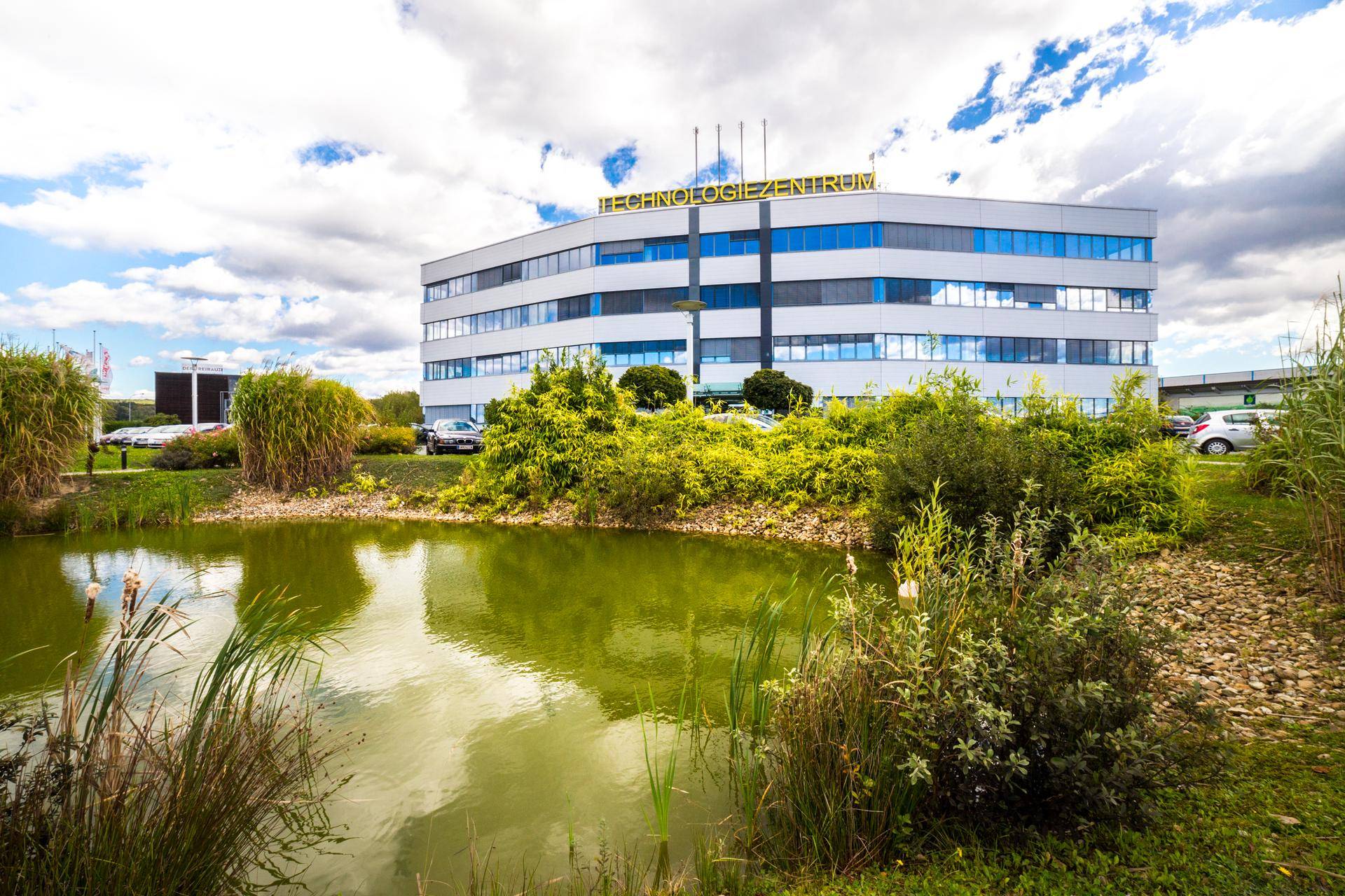 Ansicht des Technologiezentrums mit einem Teich und üppiger Grünfläche im Vordergrund unter blauem Himmel.