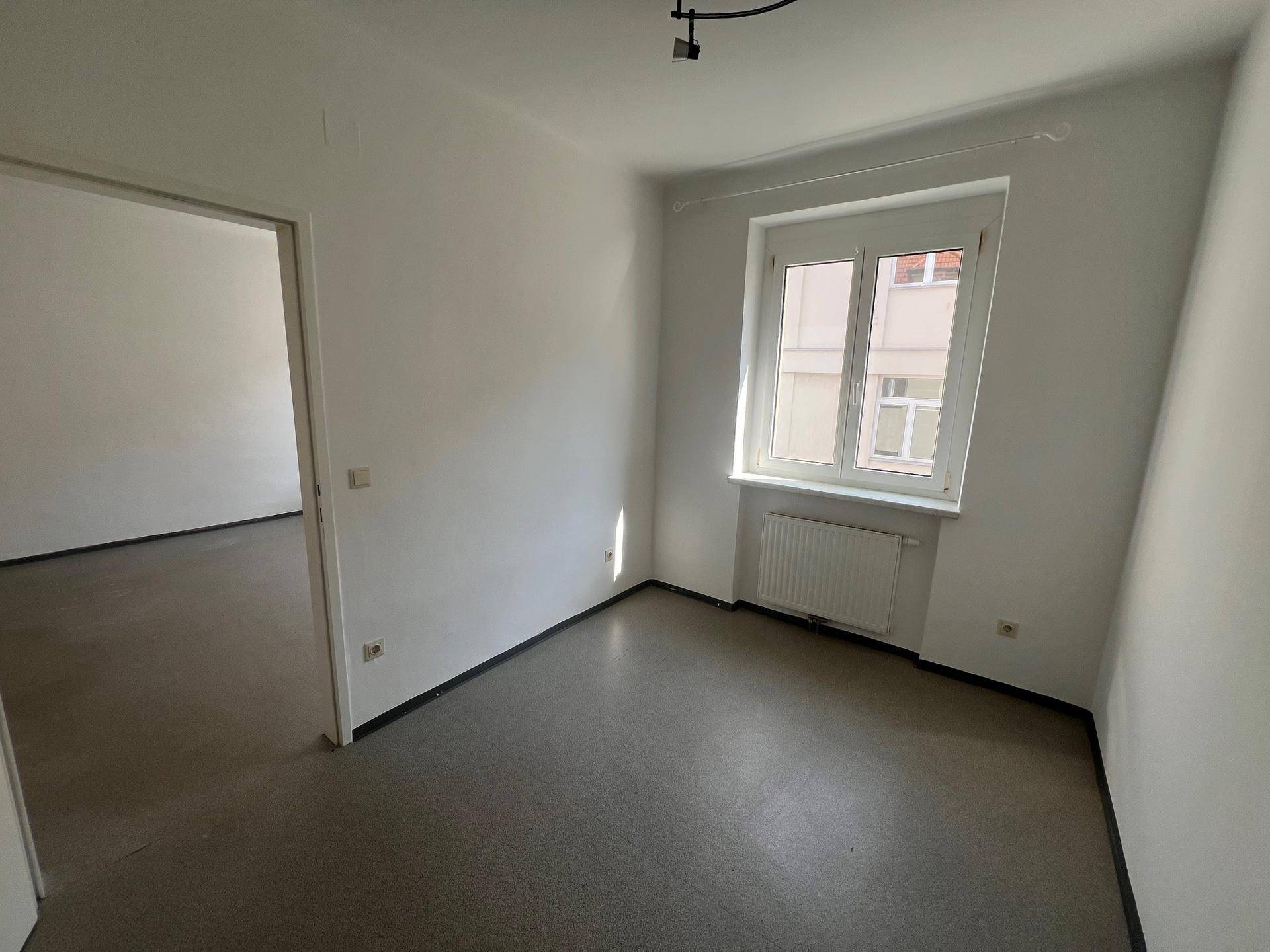 Leeres Schlafzimmer mit einem Fenster, weißen Wänden und dunklem Boden, das viel natürliches Licht erhält.