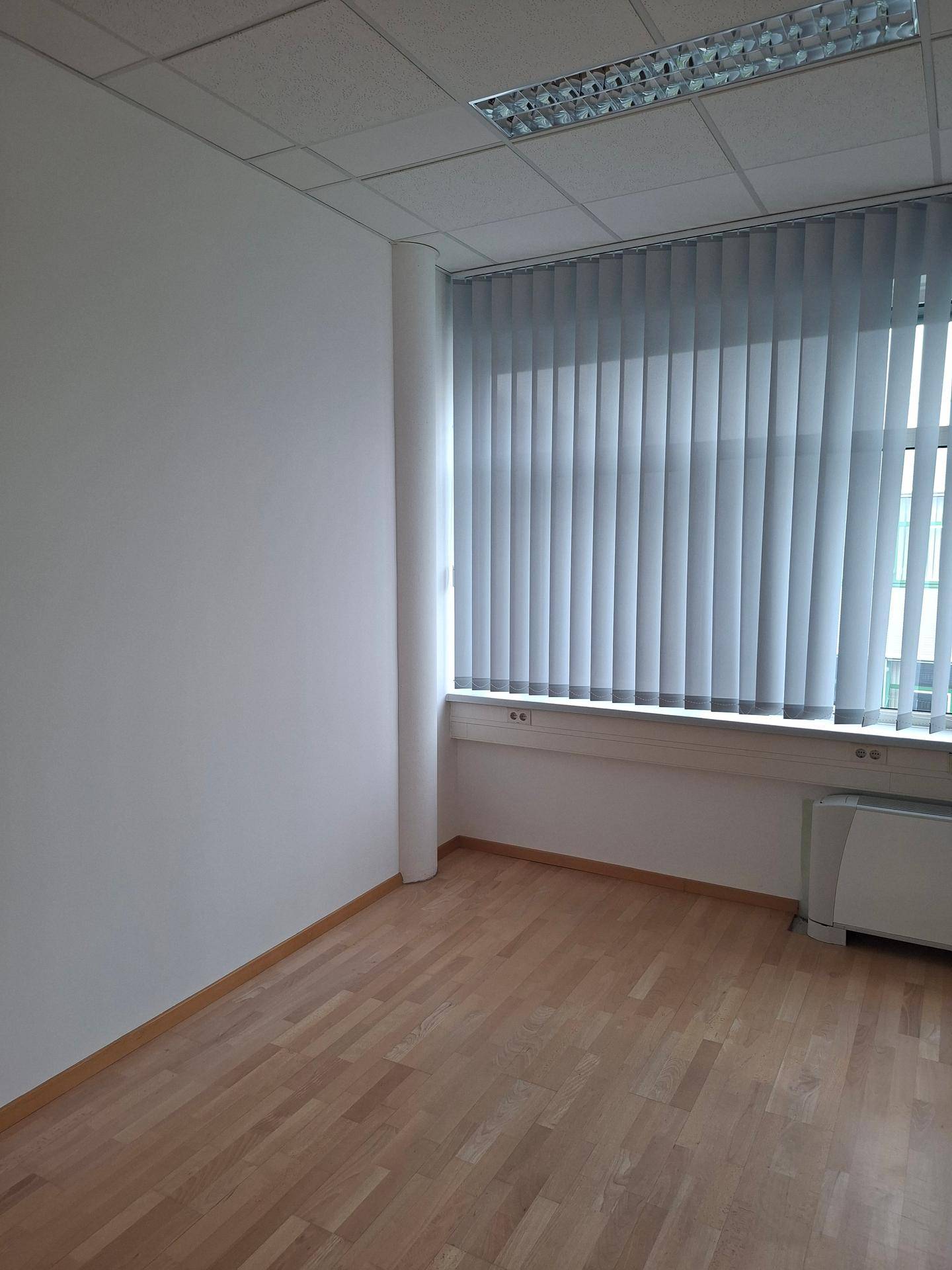 Ecke eines leeren Büros mit hellem Laminatboden, Fenster mit Jalousien und Heizkörper unter dem Fenster.