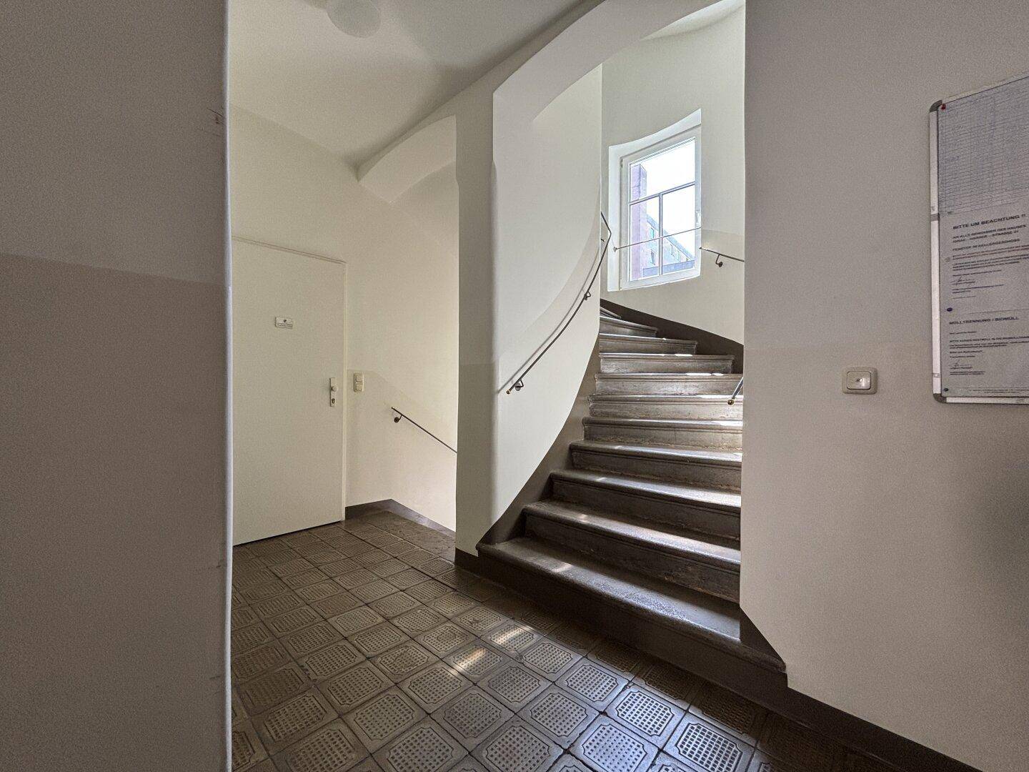 Helles Treppenhaus mit geschwungener Steintreppe und Fenster, das Tageslicht hereinlässt.