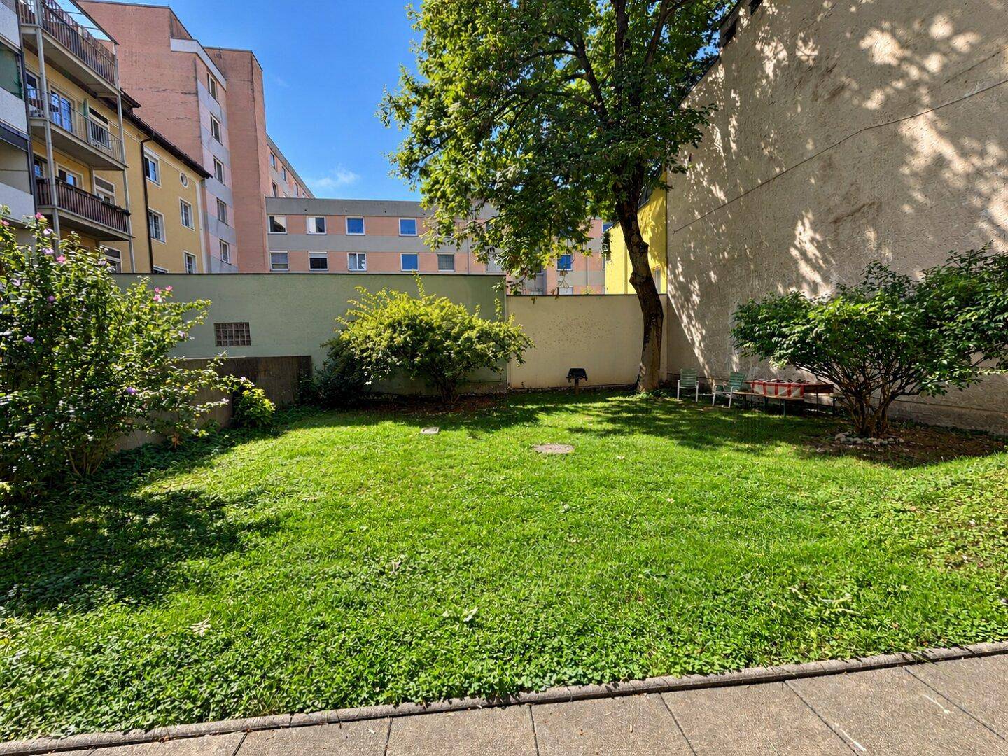 Grüner Gartenbereich mit Rasen und Bäumen, umgeben von Gebäuden in einem Innenhof.
