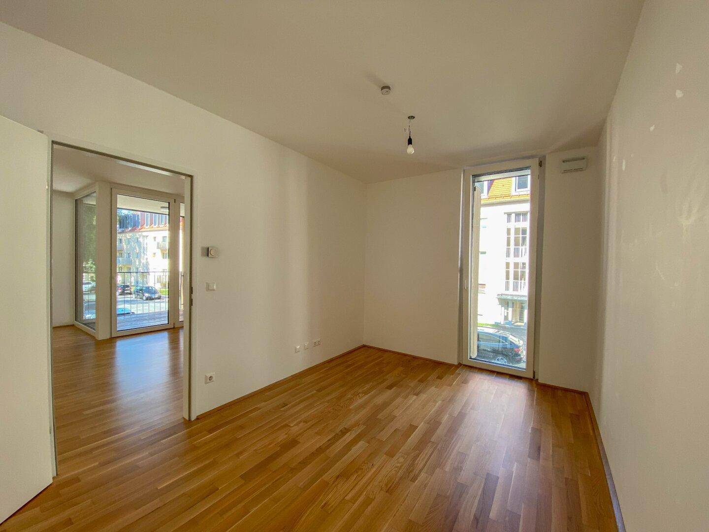 Leerer, heller Raum mit Parkettboden, großen Fenstern und Zugang zu einem weiteren Bereich mit Balkon.