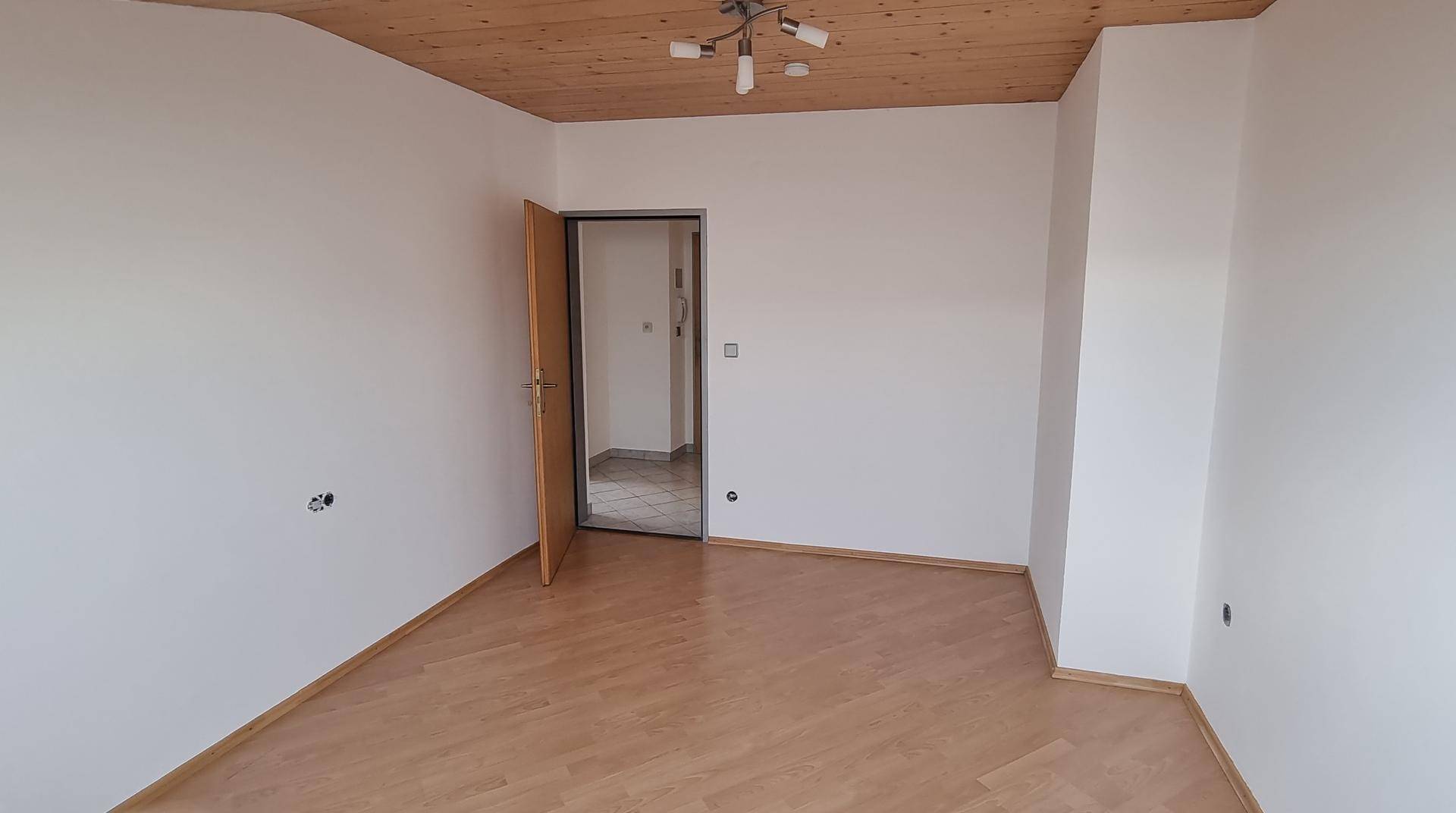 Geräumiges Zimmer mit hellem Parkettboden, weiß gestrichenen Wänden und Holztür.