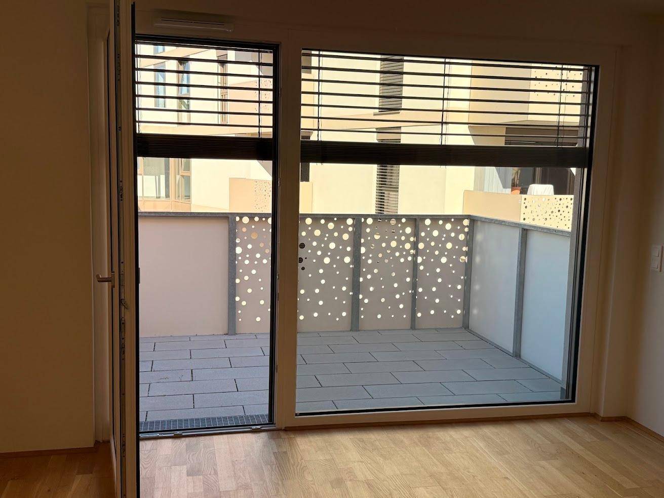 Heller Raum mit Zugang zu einem Balkon, ausgestattet mit großen Fenstern und Jalousien.