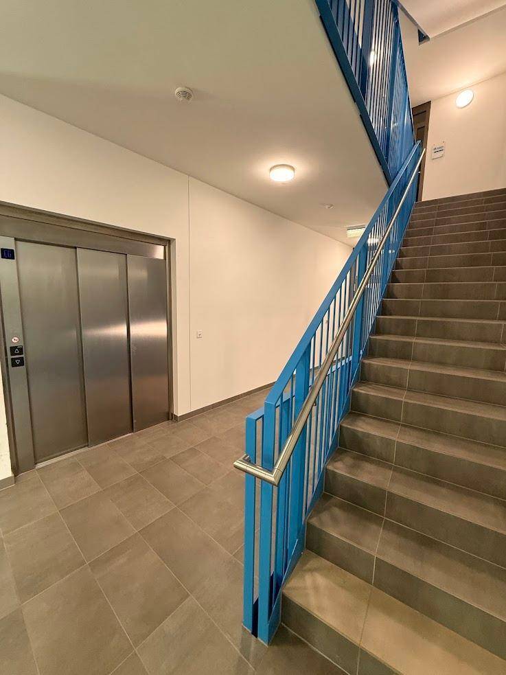 Heller Treppenaufgang mit blauen Geländern und einem modernen Aufzug in einem Wohngebäude.