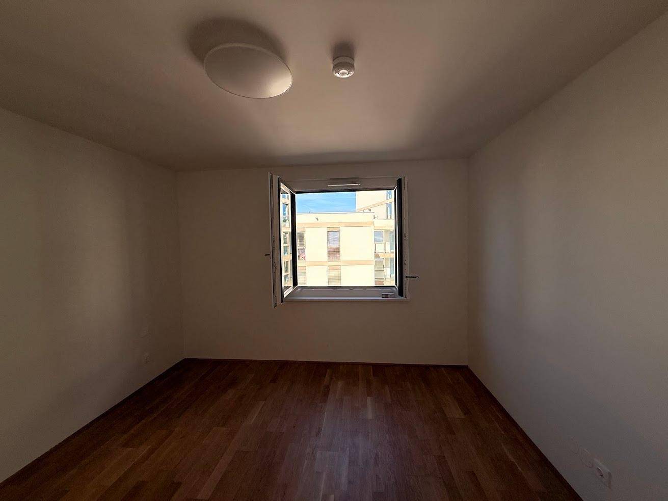 Helles Zimmer mit Parkettboden und offenem Fenster, das frische Luft hereinlässt.
