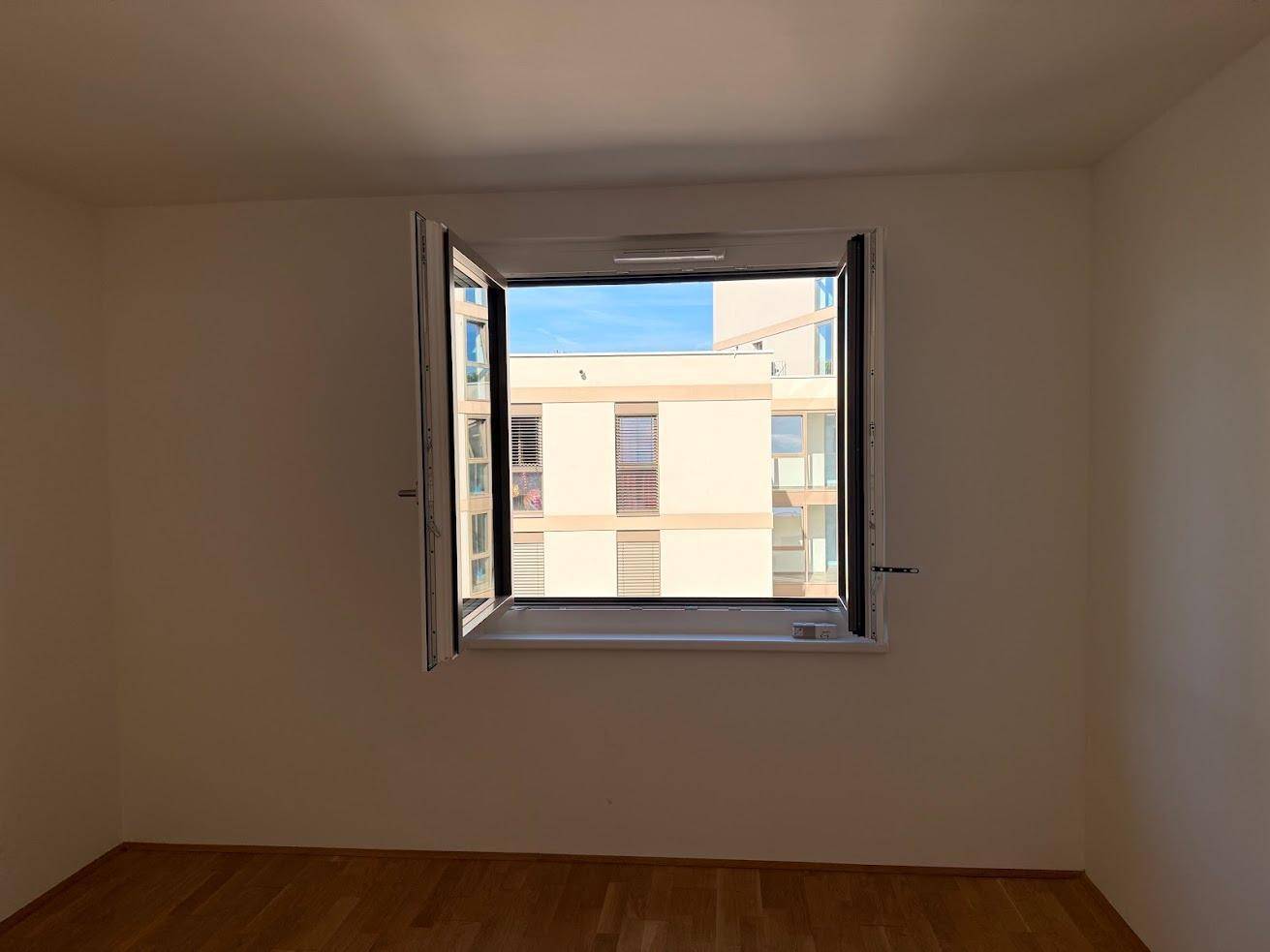Geräumiges Zimmer mit geöffnetem Fenster, Parkettboden und Blick auf die Umgebung.