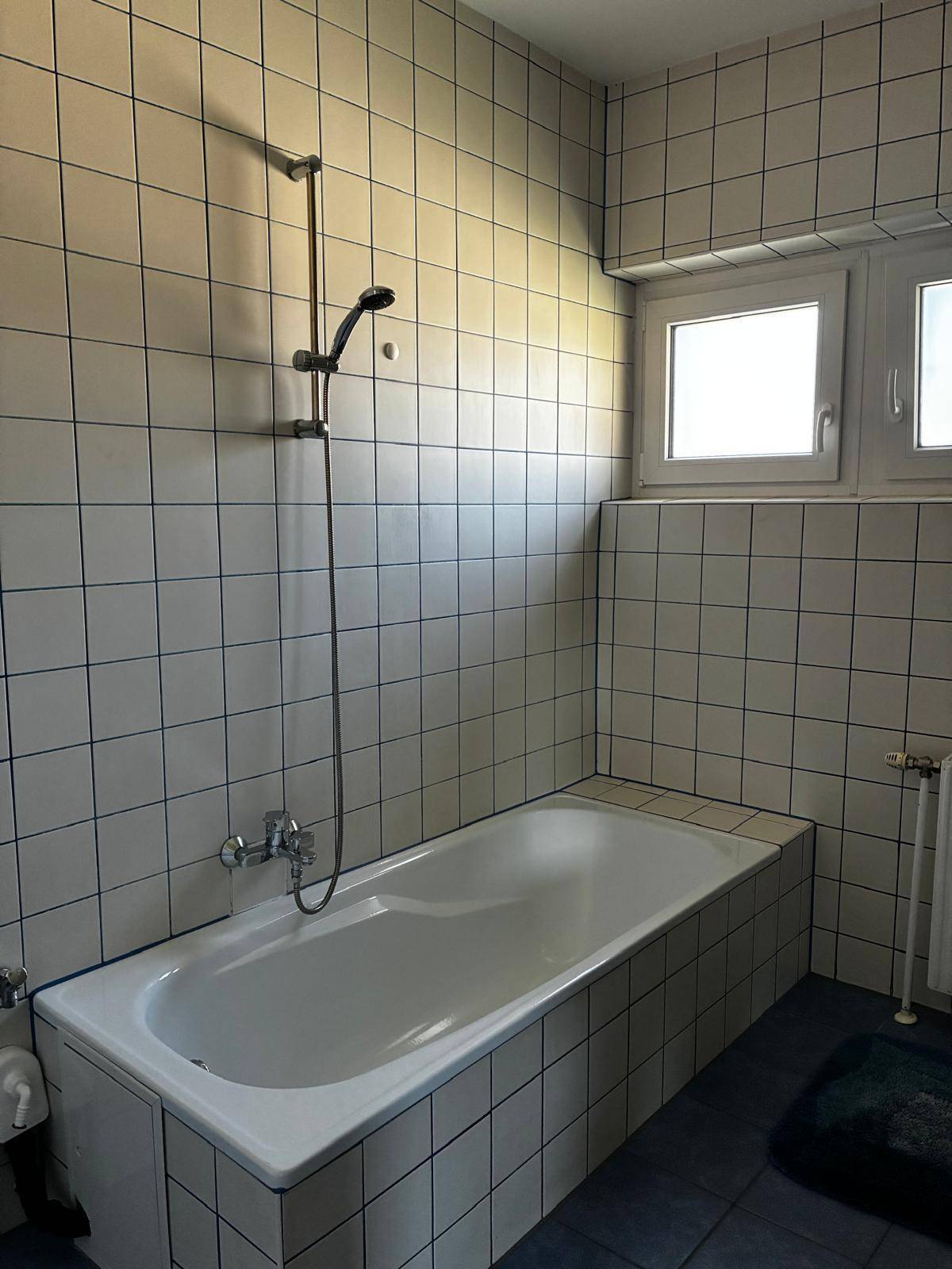 Badezimmer mit Badewanne, weißen Wandfliesen, blauen Bodenfliesen und einem Fenster.