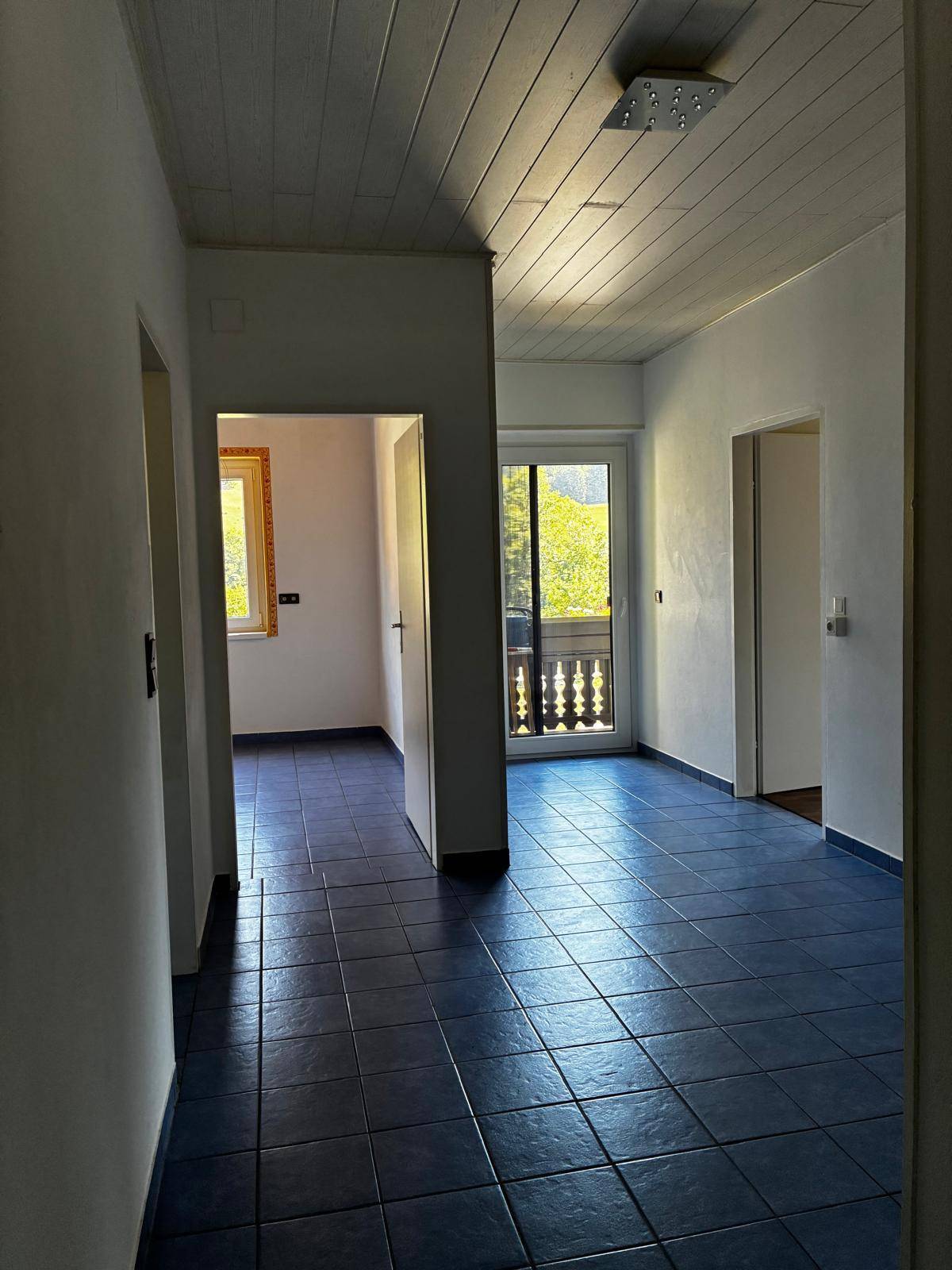 Heller Flur mit blauen Fliesen, weiß gestrichenen Wänden und Zugang zu einem Balkon.