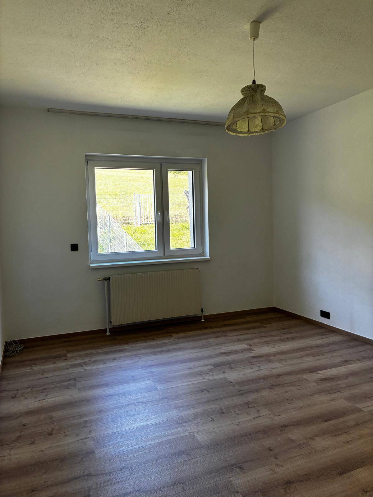 Helles, leeres Zimmer mit Laminatboden, weiß gestrichenen Wänden und Blick ins Grüne.