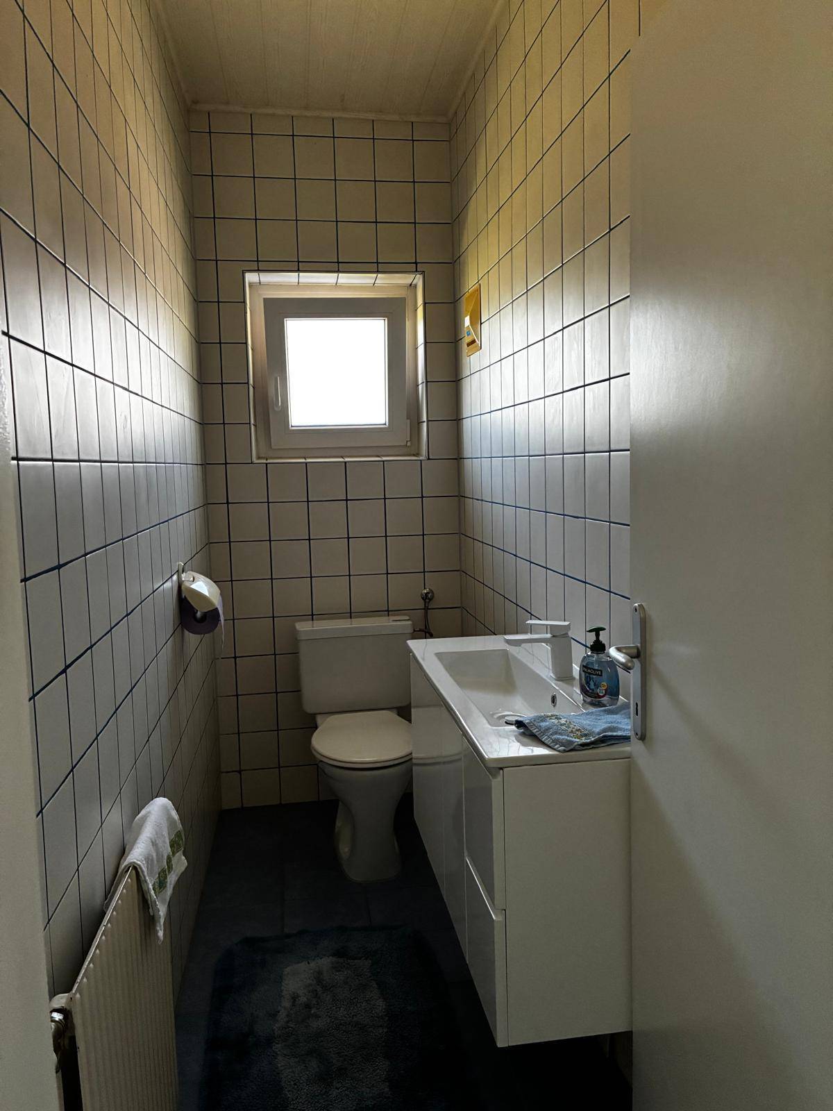 Kleines WC mit Fenster, weißen Fliesen, blauen Bodenfliesen und einem wandhängenden Waschbecken.