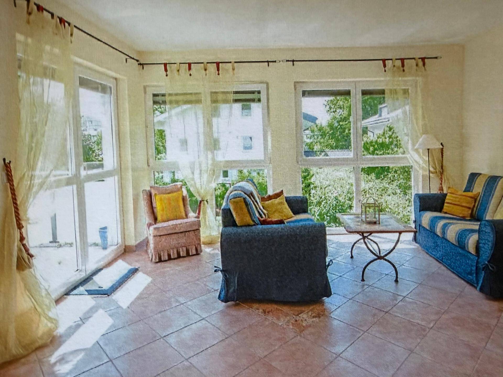 Helles Wohnzimmer mit großen Fenstern, Fliesenboden und Zugang zum Außenbereich.