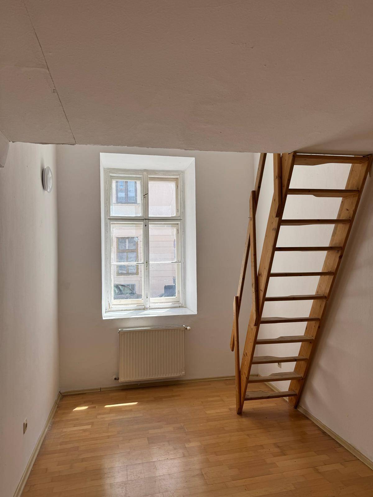 Heller Raum mit Holzboden, Fenster und einer Holztreppe, die in eine obere Etage führt.