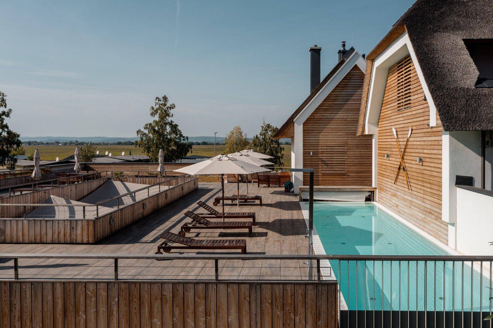 Außenbereich mit Swimmingpool, Holzdeck und Liegestühlen unter Sonnenschirmen.
