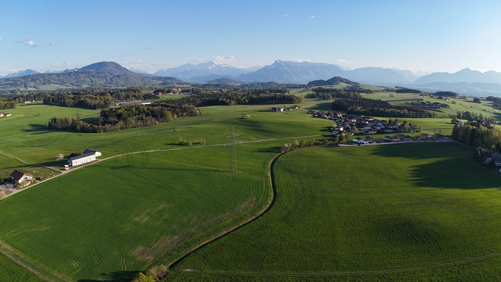 Weitläufige grüne Landschaft mit Feldern, Wäldern und einer Bergkette im Hintergrund unter blauem Himmel.