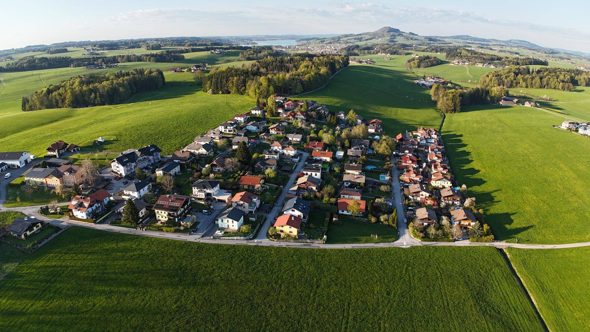Luftaufnahme einer Wohnsiedlung eingebettet in weite grüne Felder mit bewaldeten Hügeln im Hintergrund.
