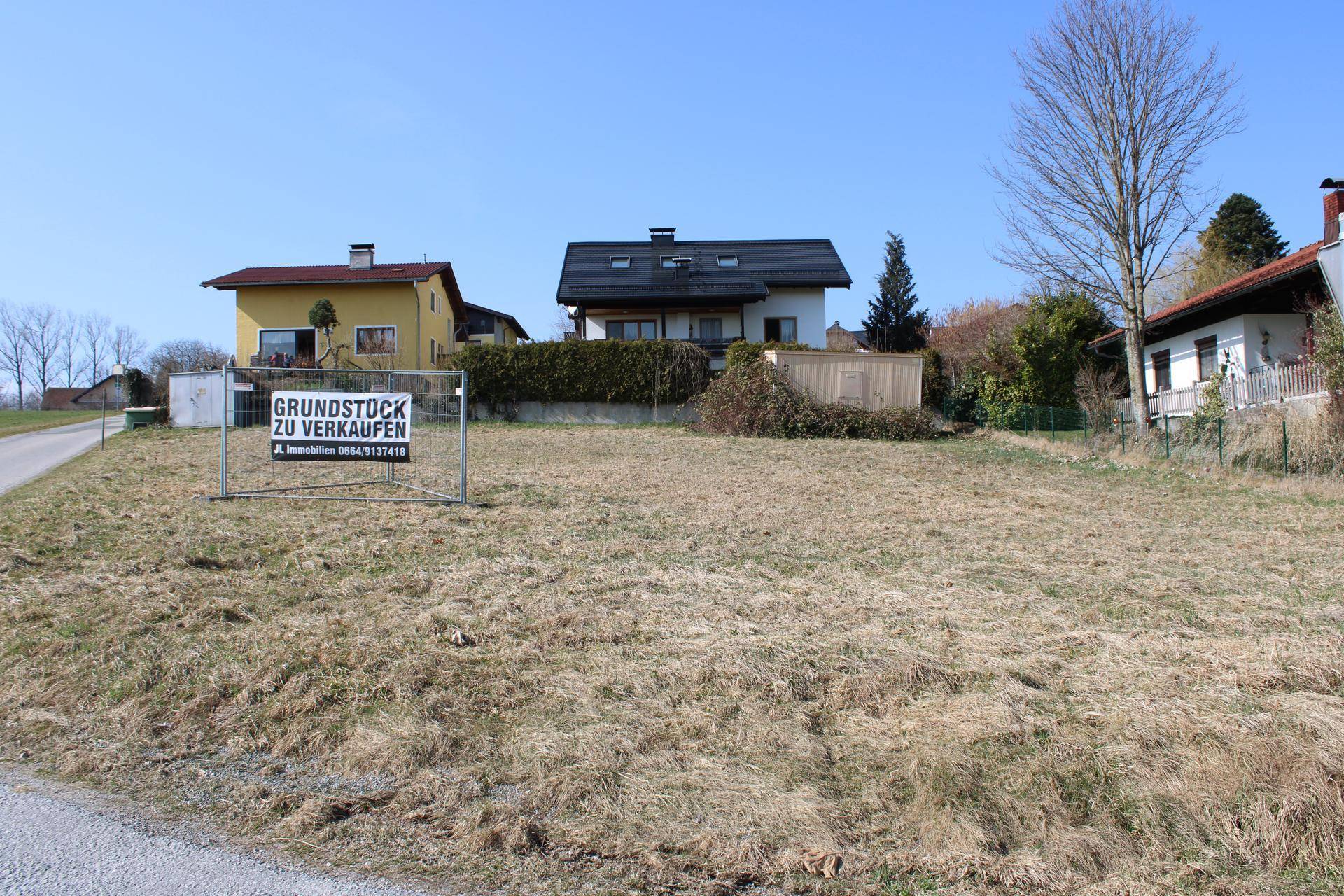 Grundstück zum Verkauf mit angrenzenden Wohnhäusern und ländlicher Umgebung unter blauem Himmel.