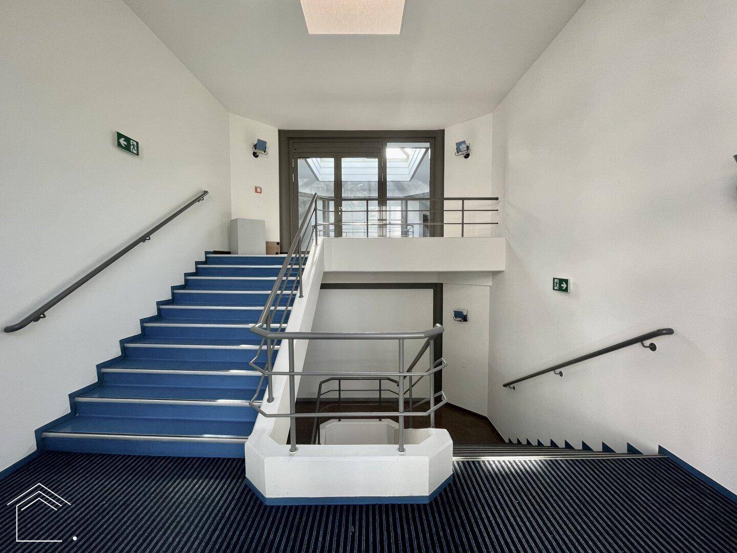 Helles Treppenhaus mit blauen Stufen und Metallgeländer, das zu verschiedenen Etagen führt.