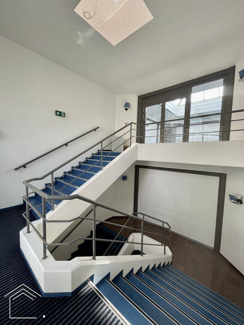 Helles Treppenhaus mit blauen Stufen und Metallgeländer, das zu verschiedenen Etagen führt.