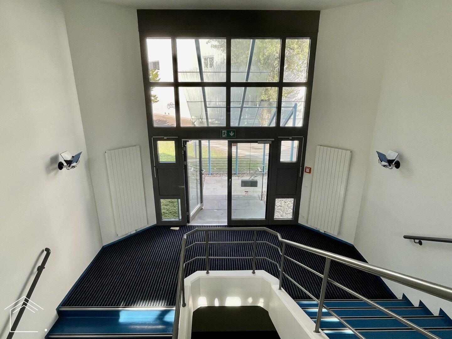 Heller Eingangsbereich mit großer Glasfront und Treppe, bietet Zugang zum Gebäude.