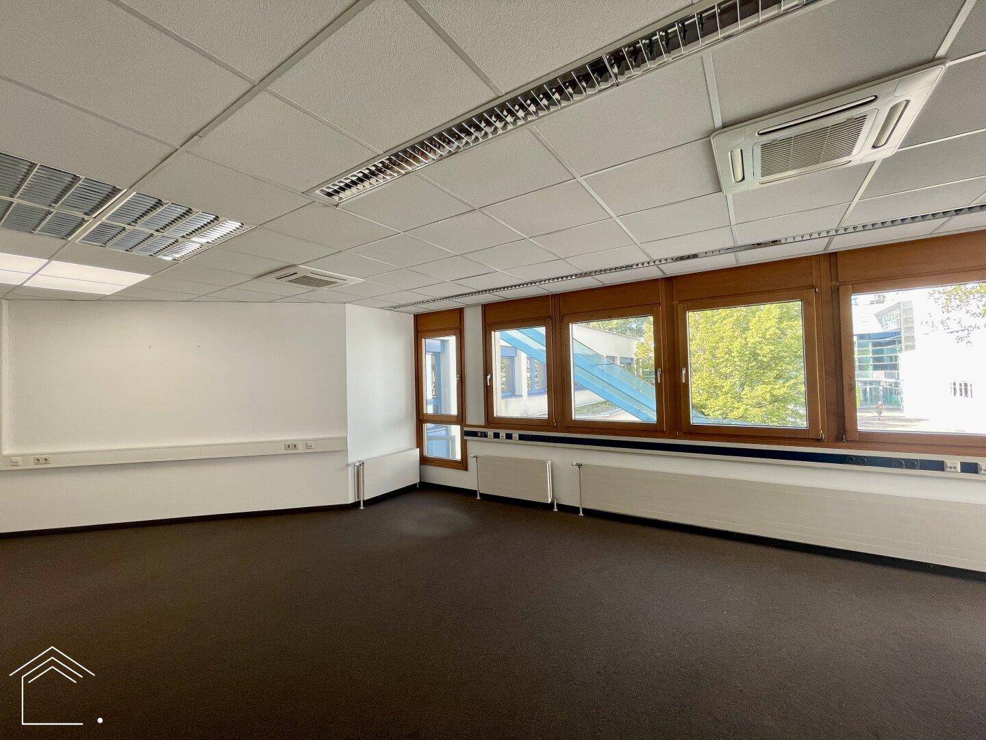 Geräumiges Büro mit Teppichboden, großen Fenstern und Klimaanlage, ideal für produktives Arbeiten.