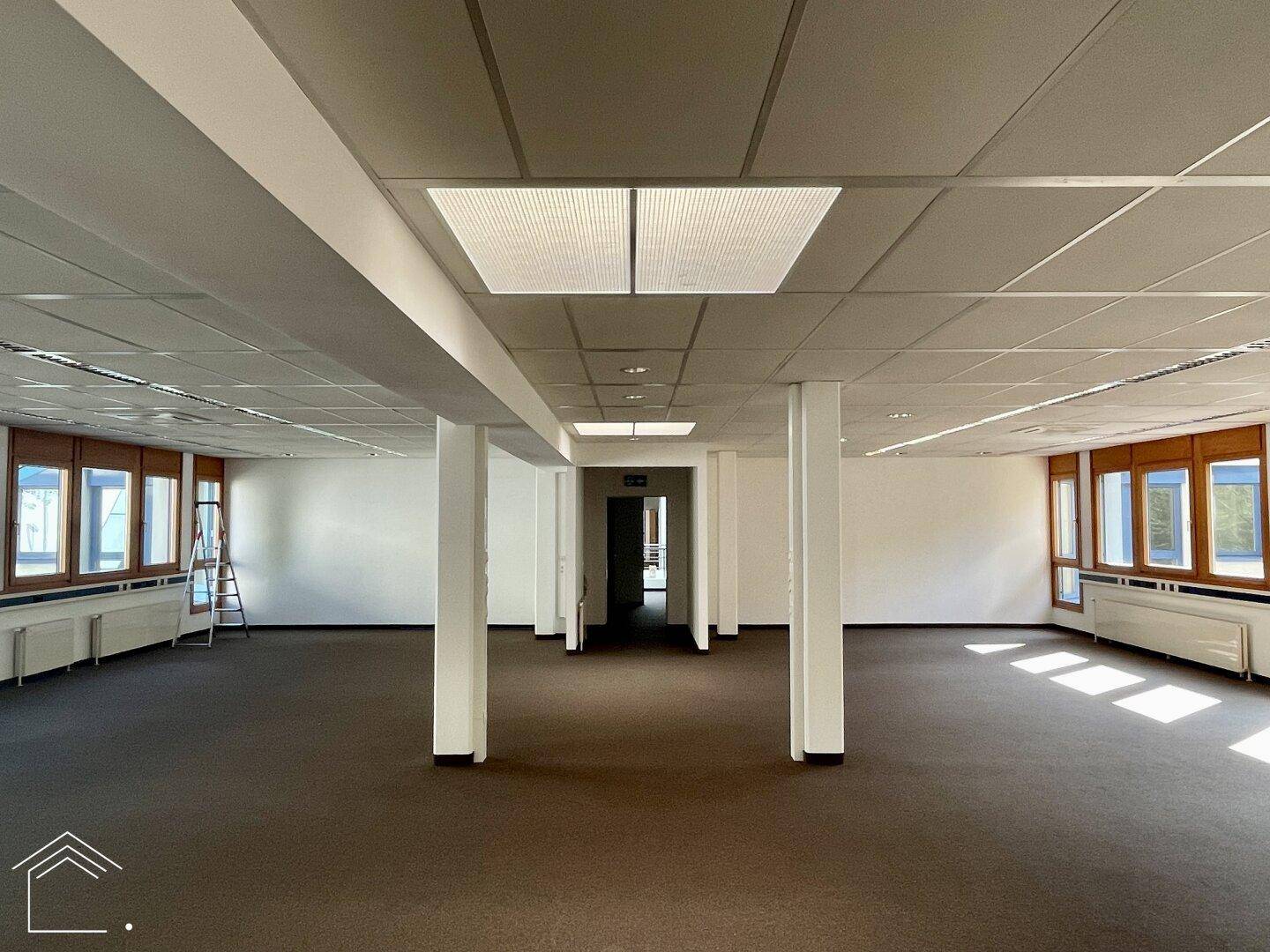 Weitläufiges Büro mit Teppichboden, Säulen und Deckenbeleuchtung, ideal für Großraumbüros.