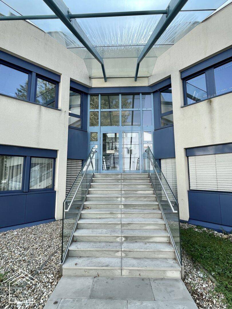 Moderner Gebäudeeingang mit Glasfassade und Treppe, umgeben von Kies und Grünflächen.