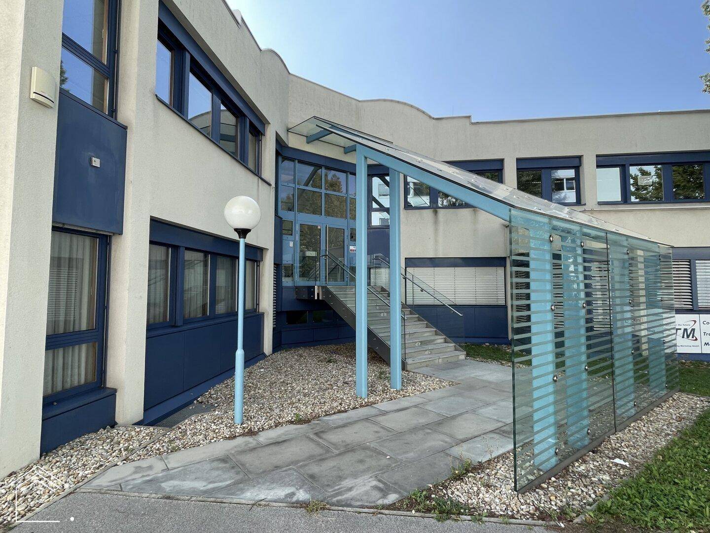 Moderne Gebäudefassade mit Glasüberdachung und blauen Akzenten, umgeben von Grünflächen.