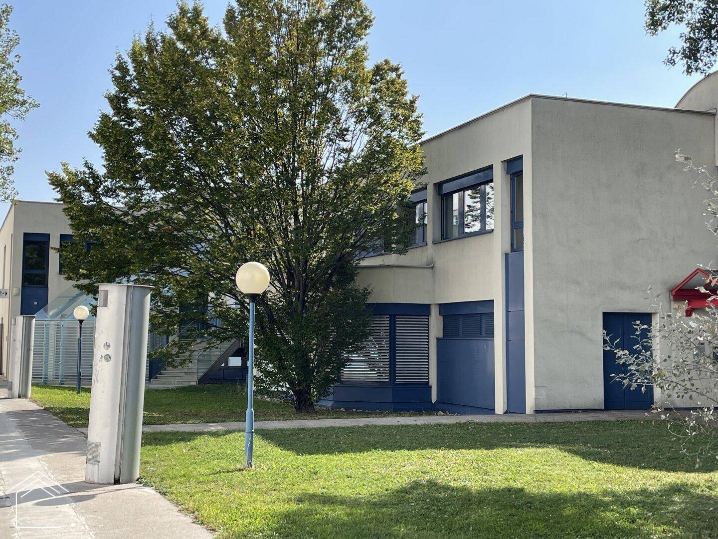 Moderne Gebäudefassade mit blauen Akzenten und gepflegter Grünanlage, umgeben von Bäumen.
