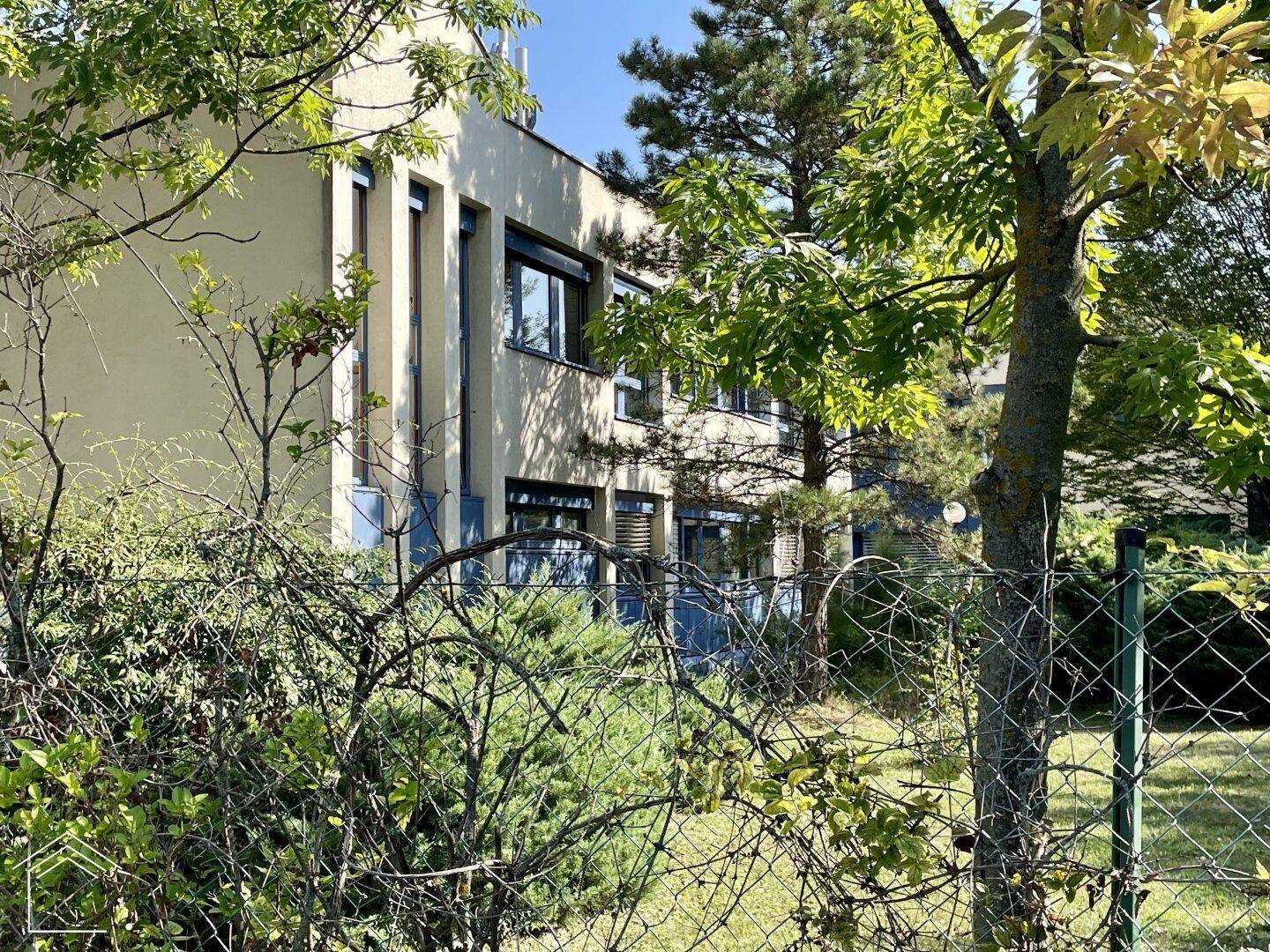 Gebäudefassade durch einen Zaun und Bäume sichtbar, mit blauen Fensterrahmen.