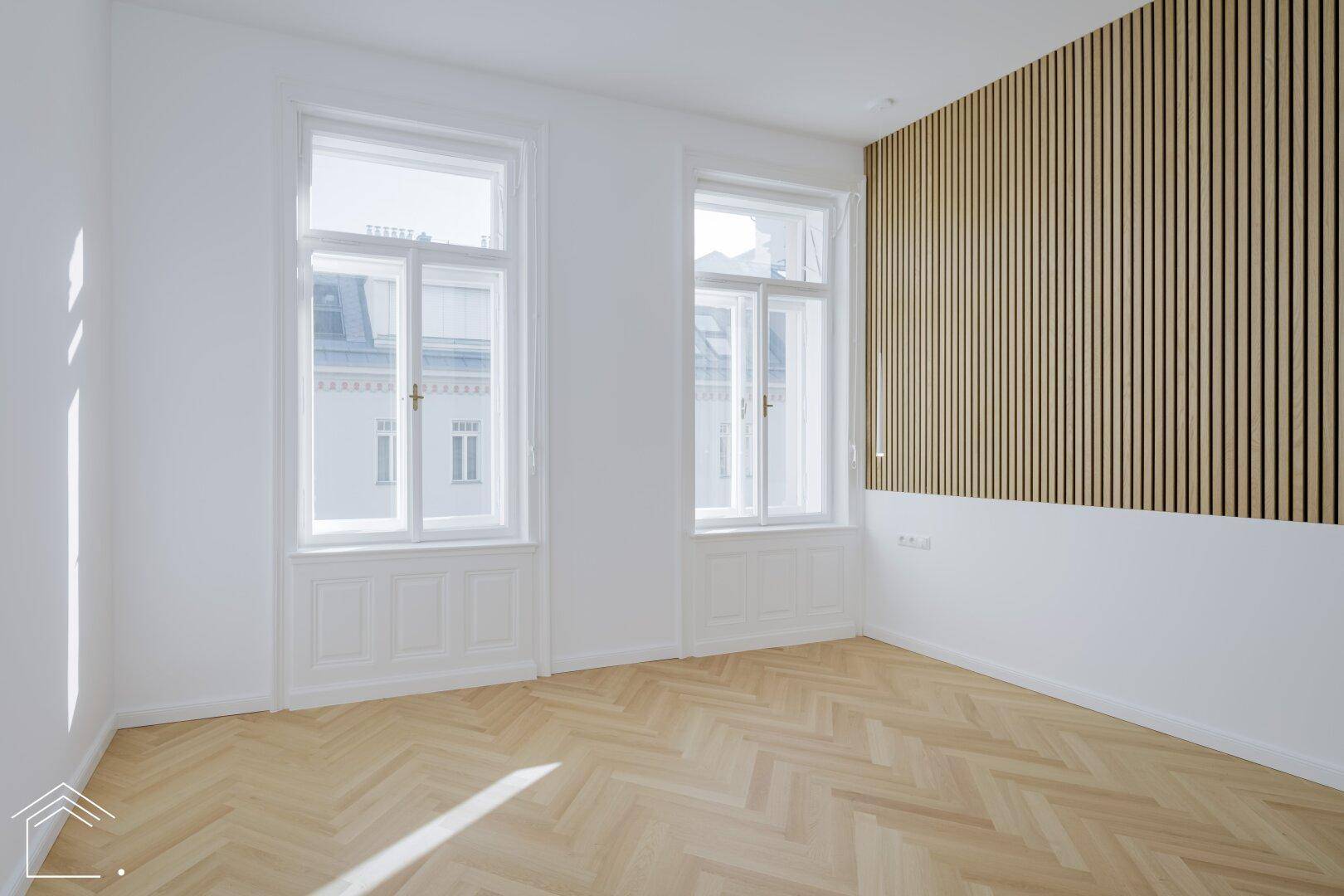 Heller Raum mit großen Fenstern, Fischgrätparkett und dekorativer Holzvertäfelung.