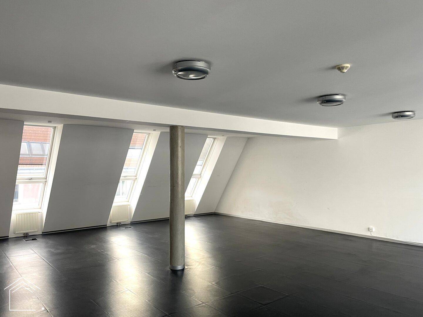 Heller Büroraum im Dachgeschoss mit schrägen Fenstern, dunklem Boden und einer zentralen Betonsäule.