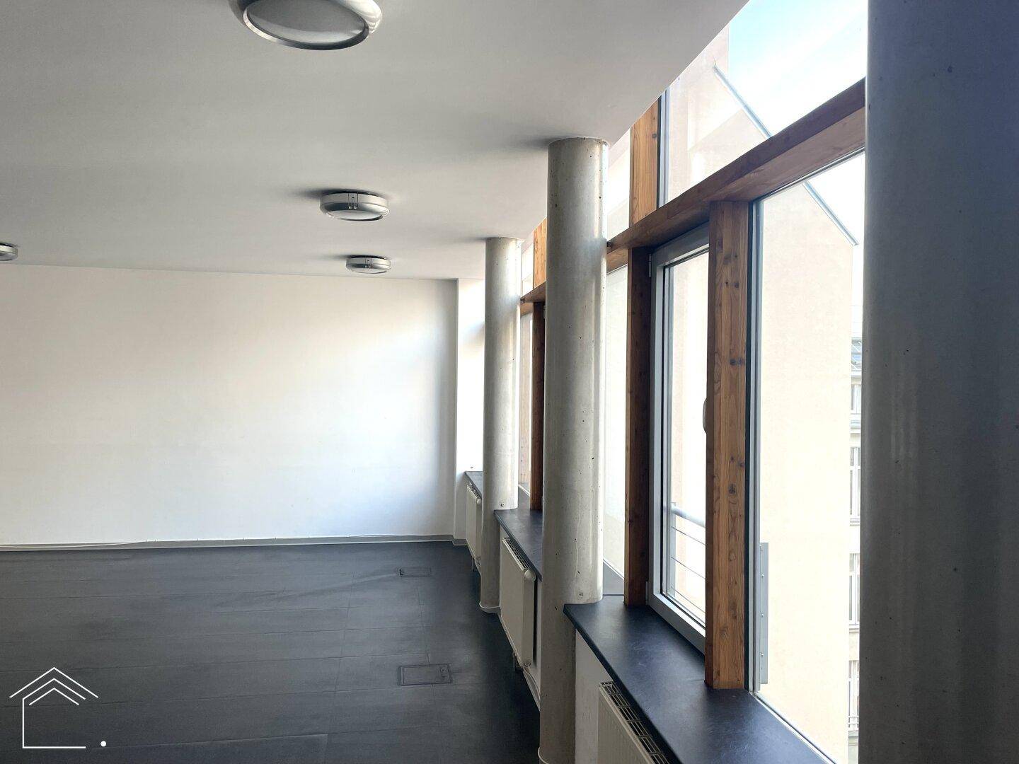Langer Flur oder offener Bürobereich mit durchgehender Fensterfront, die viel Tageslicht hereinlässt, und mehreren Betonsäulen.