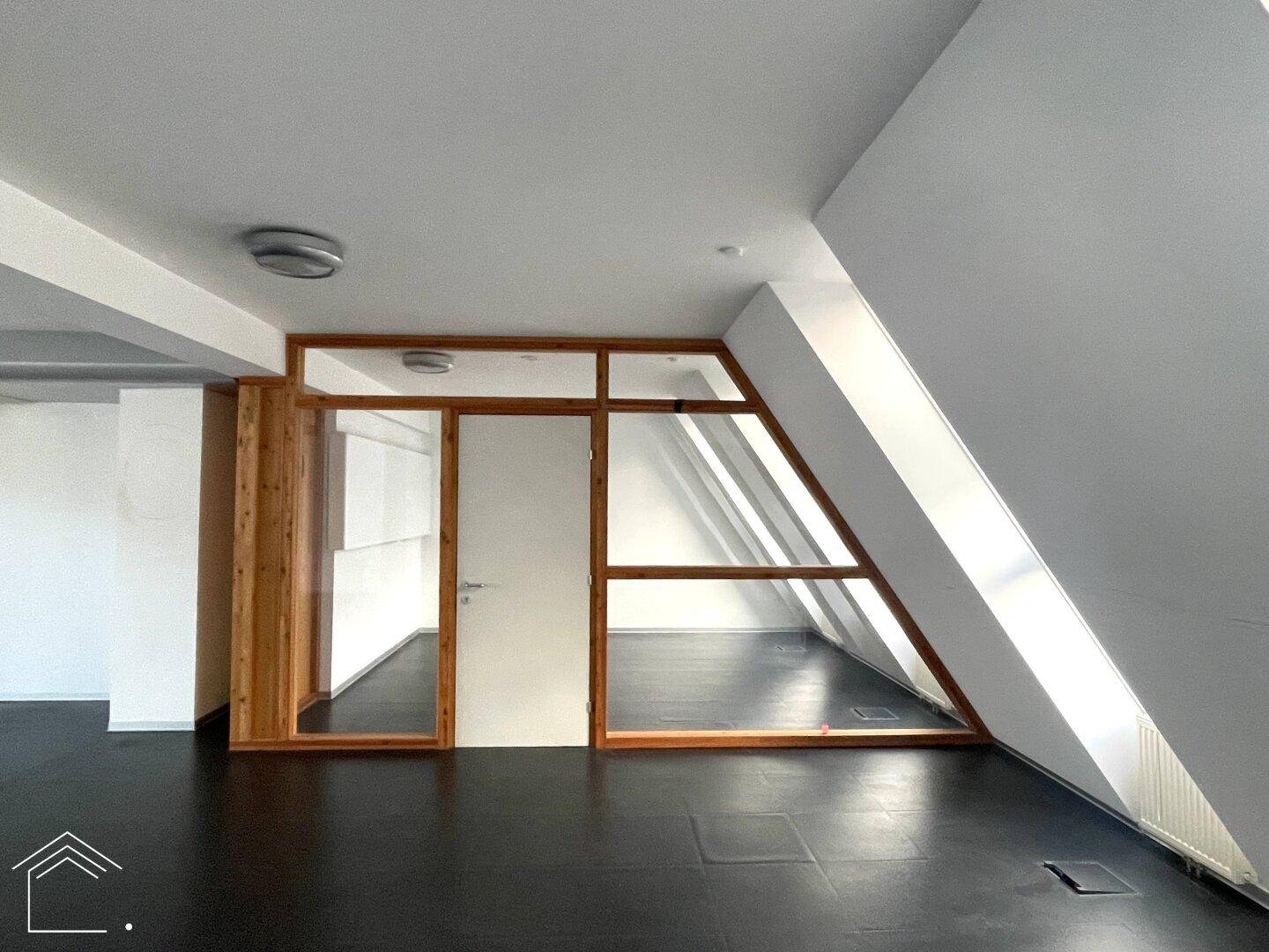 Moderner Büroraum mit schrägen Wänden, Holzelementen und einer Glasabtrennung zu einem weiteren Bereich.