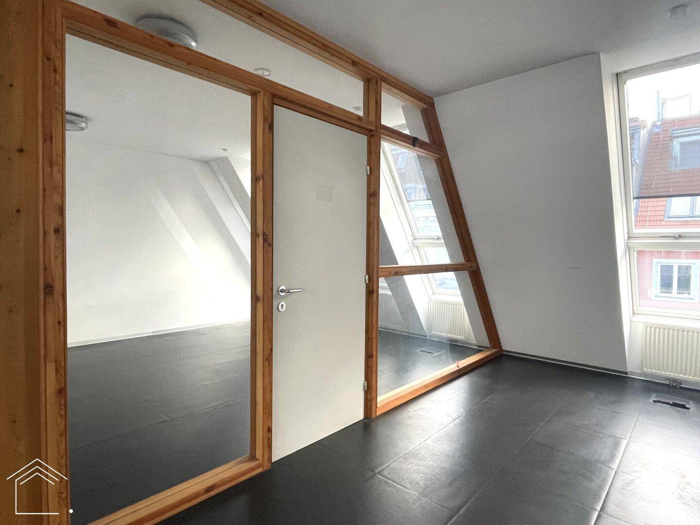 Heller Büroraum mit schrägen Dachfenstern, Holzelementen und einer Glastür zu einem Nebenraum.
