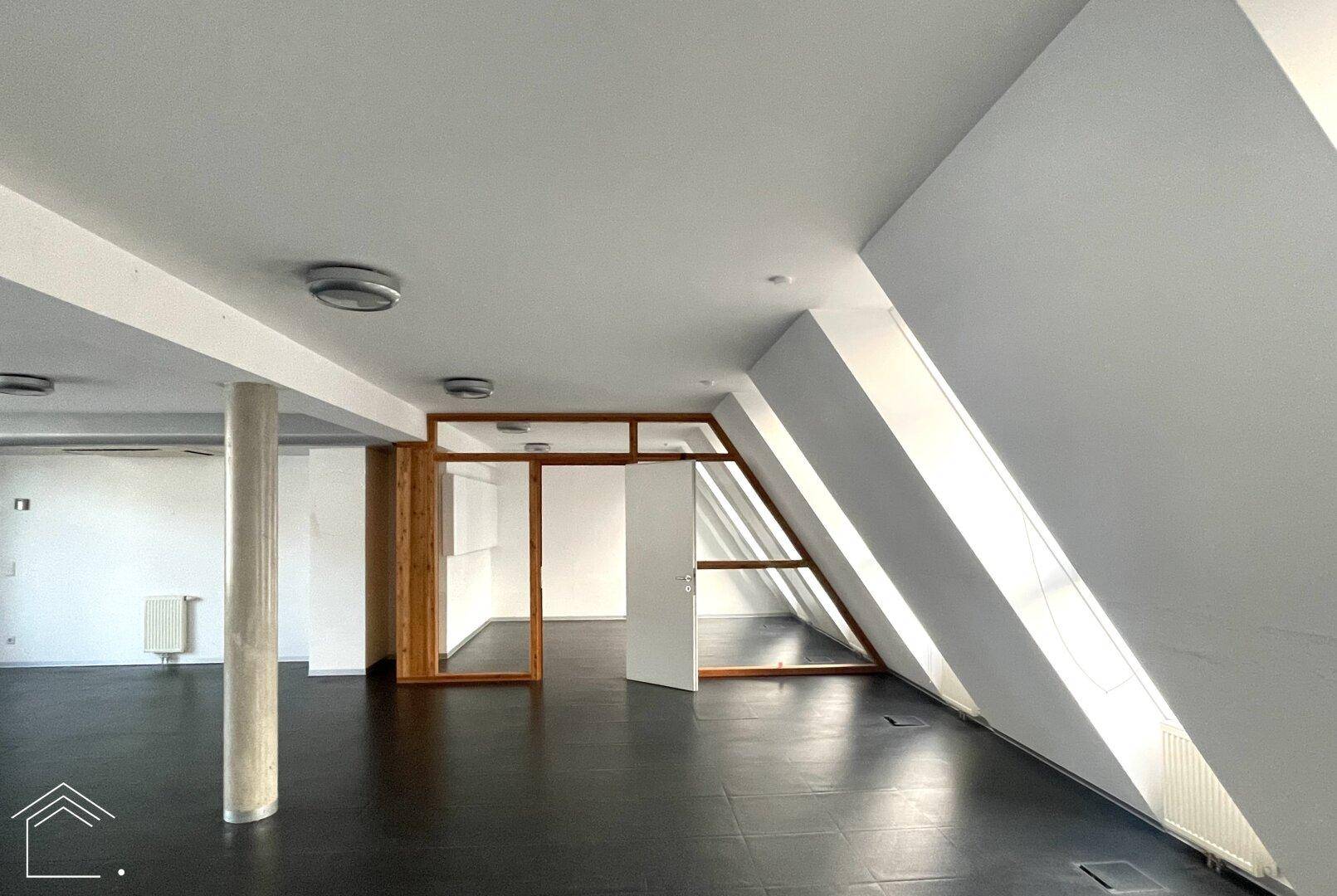Großer, heller Büroraum mit dunklem Boden, schrägen Dachfenstern und einer zentralen Betonsäule.